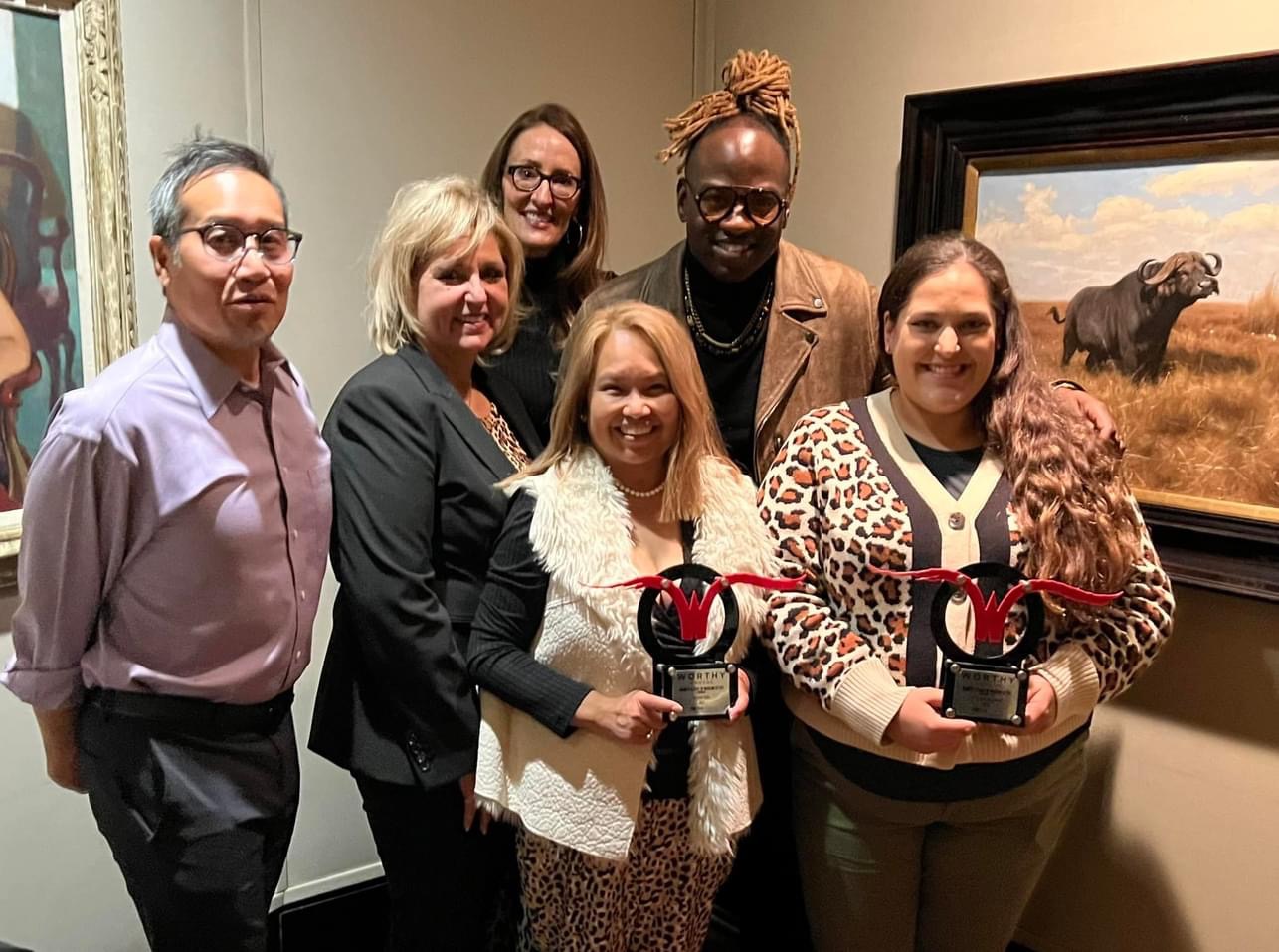 The Office of Communications and Strategy poses with two "Worthy" awards from the Greater Fort Worth PRSA. They are posed in a corner in front of two pieces of art at the Fort Worth Zoo.