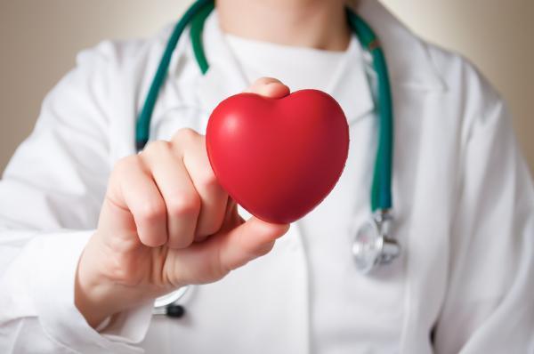 A person (no face shown) is wearing a white coat and a teal stethoscope while holding a red heart in between their index finger and thumb in their right hand that is held in the middle of the frame.